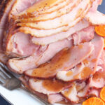 side shot of sliced bourbon glazed ham on a white platter with sliced oranges on the side