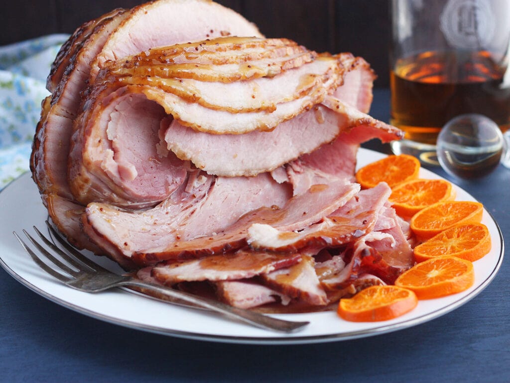 side shot of sliced bourbon glazed ham on a white platter with sliced oranges on the side and a bottle of bourbon in the background