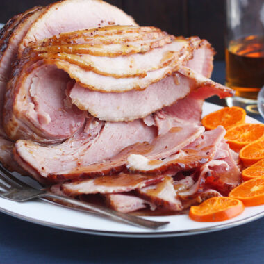 side shot of sliced bourbon glazed ham on a white platter with sliced oranges on the side and a bottle of bourbon in the background