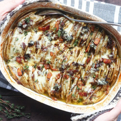 Overhead shot of a pair of hands placing a white oval casserole dish with baked scalloped potatoes with bacon on a black and white napkin.