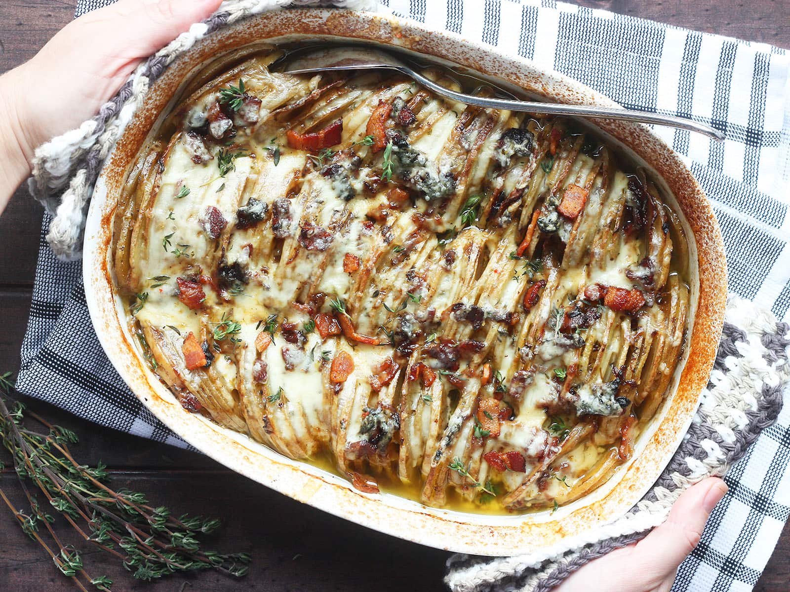 overhead shot of a pair of hands placing a white oval casserole dish with baked scalloped potatoes with bacon on a black and white napkin