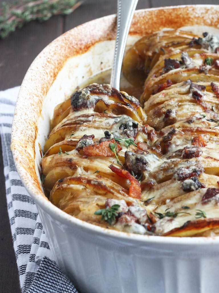 Metal spoon scooping out a serving of scalloped potatoes with bacon from a white oval casserole dish.