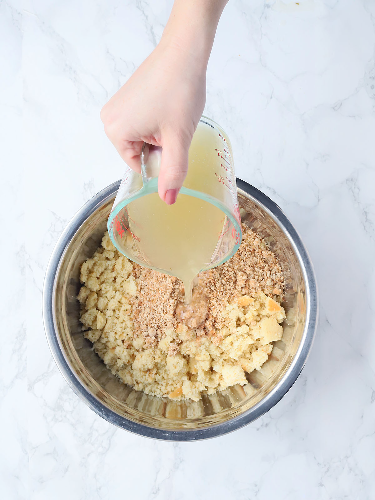 hand pouring chicken broth into a mixing bowl of crumbled cornbread and breadcrumbs