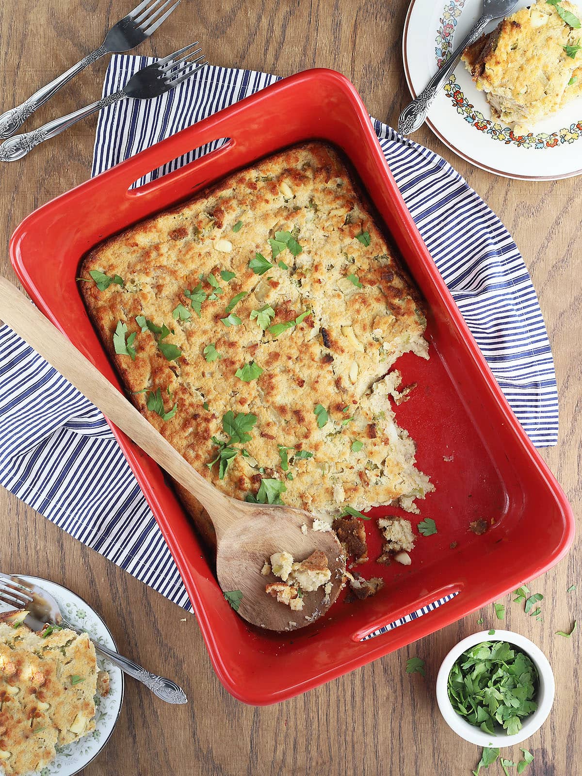 Large wooden spoon serving southern cornbread dressing with chicken from a red casserole dish, plates of dressing, a bowl of chopped parsley and forks to the side.