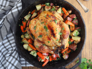 overhead shot of cast iron roast chicken with roasted vegetables, black and white dish towel, meat fork and carving knife to the side