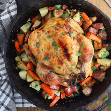 overhead shot of cast iron roast chicken with roasted vegetables, black and white dish towel, meat fork and carving knife to the side