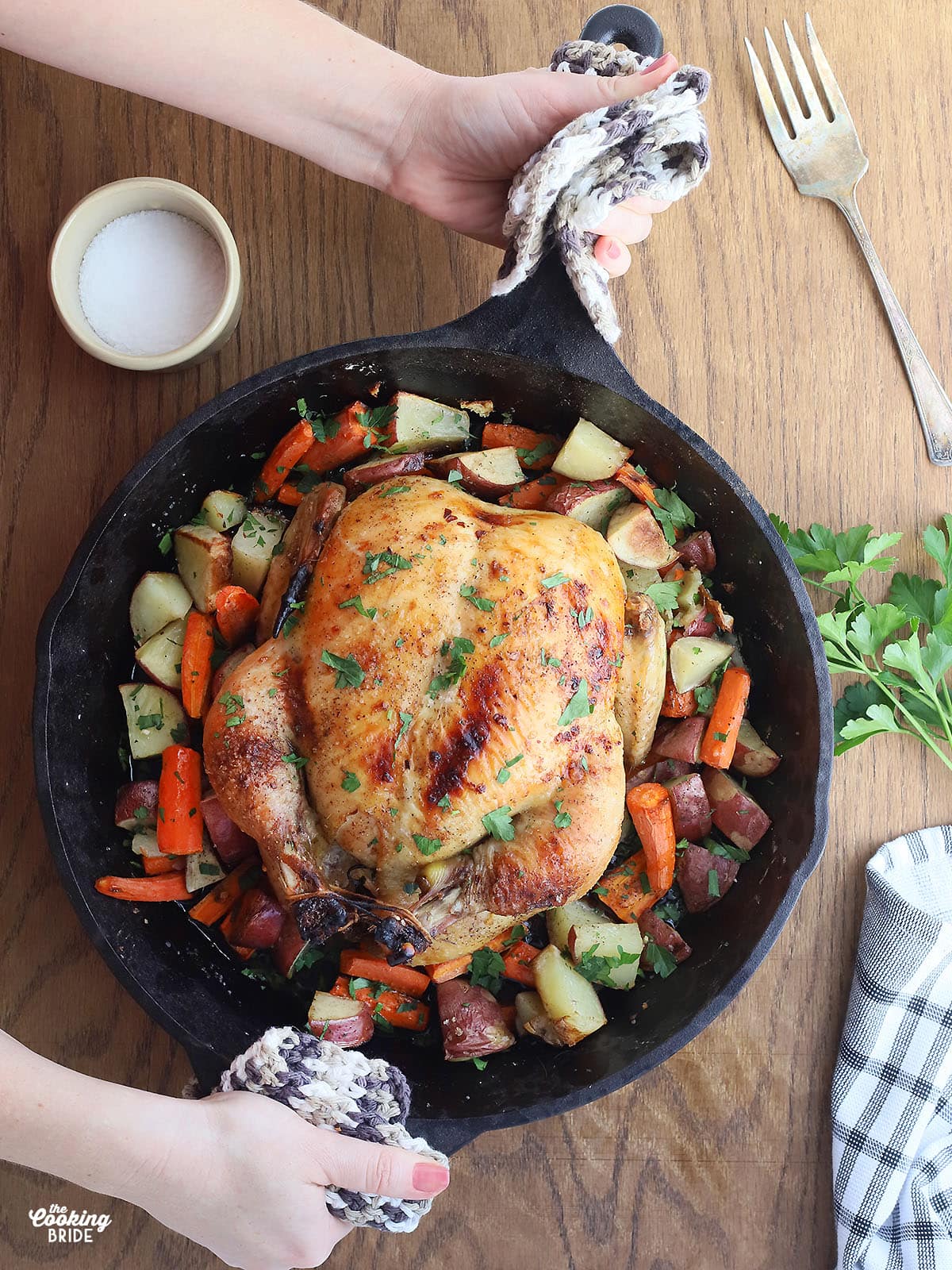 hands placing a cast iron skillet of roasted vegetables and whole chicken onto a table