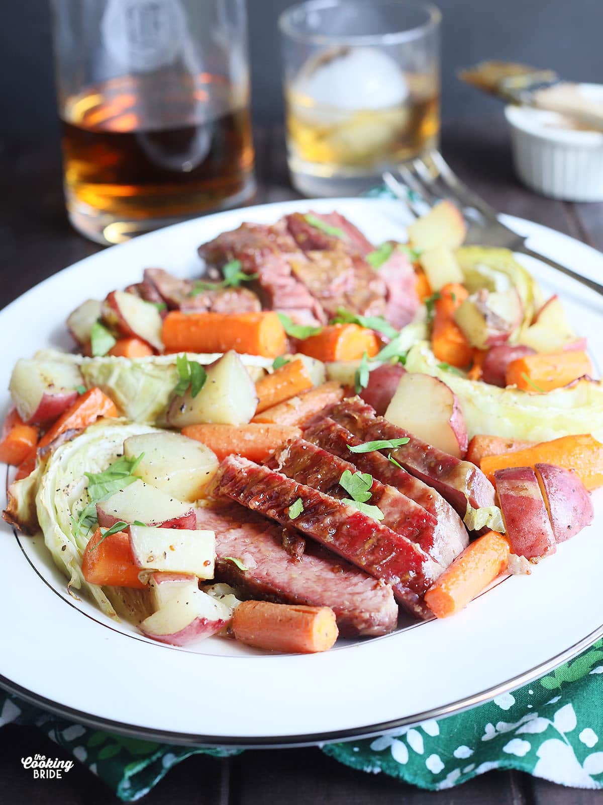 side view of sliced bourbon glazed corned beef and roasted vegetables on a white platter with a bottle of bourbon and glass in the background