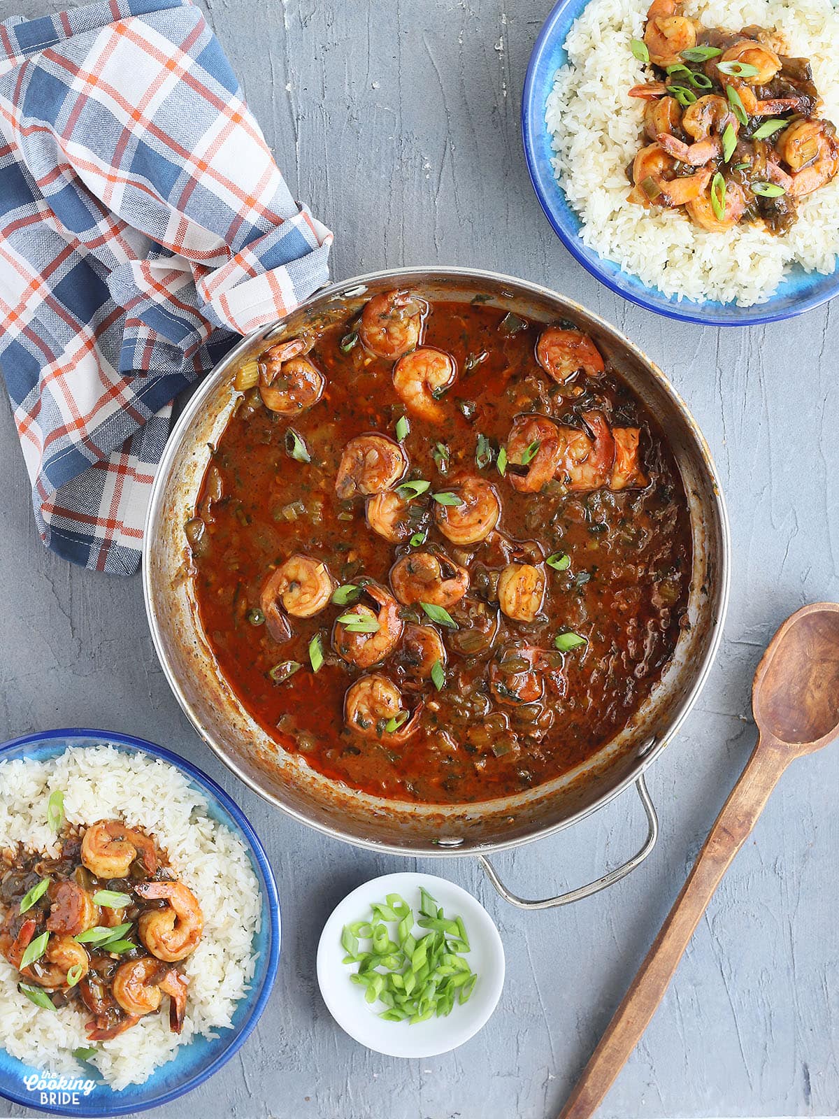 metal saucepan with cooked shrimp in gravy, two blue bowls of Cajun shrimp and rice garnished with green onions and a wooden serving spoon to the side