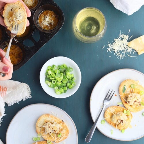Two gray plates with oysters on toast sprinkled with Parmesan cheese, sliced green onions, glasses of white wine, a hand spooning an oyster onto a piece of bread.