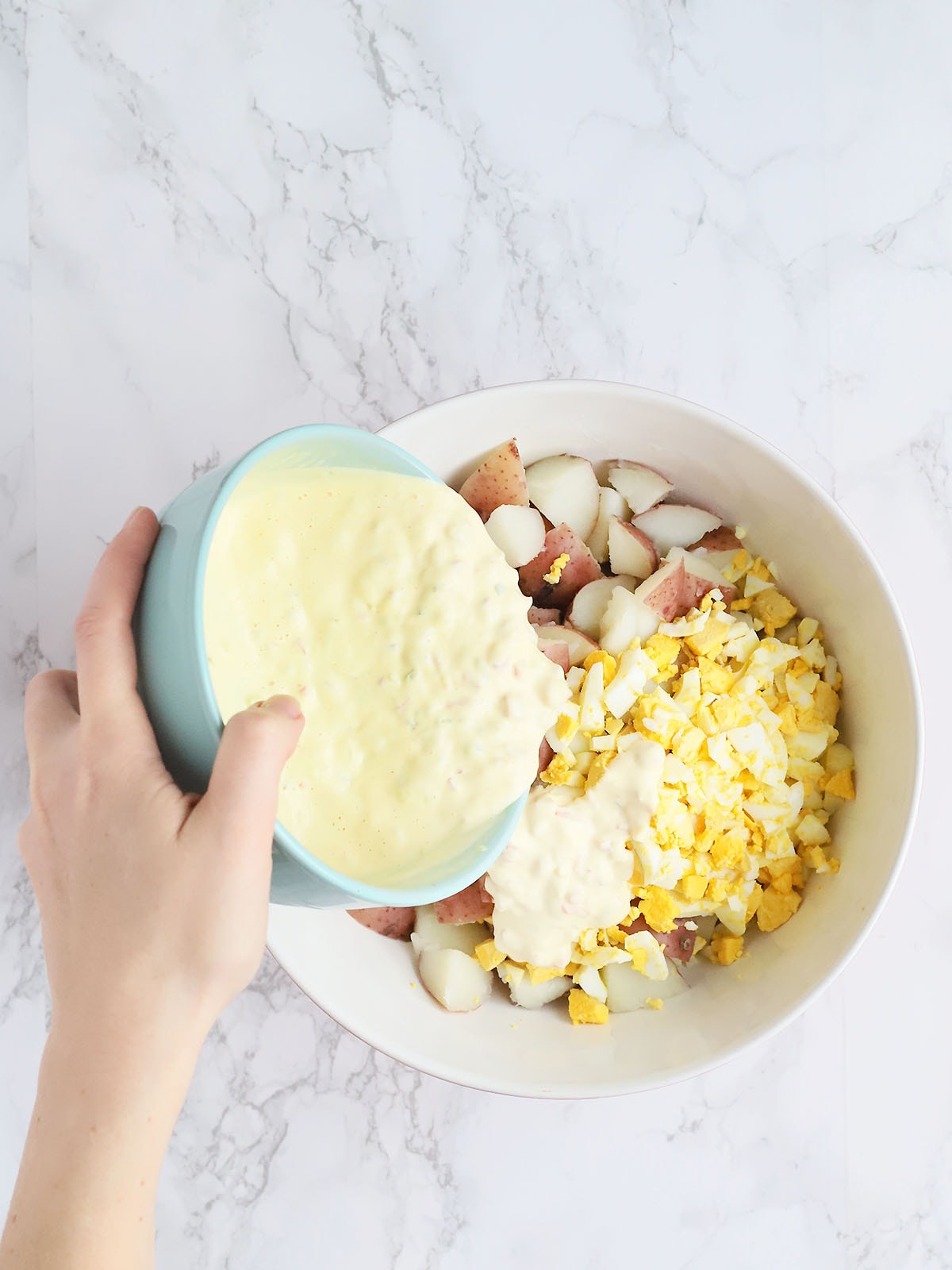 hand pouring mayonnaise mixture over a mixing bowl of potatoes, hardboiled eggs and celery