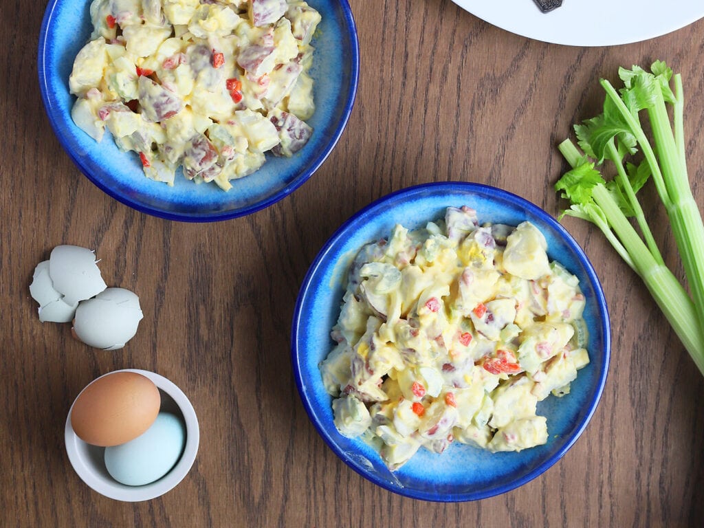 overhead shot of two blue bowls filled with potato salad, celery stalks, colorful hardboiled eggs and roast chicken to the side