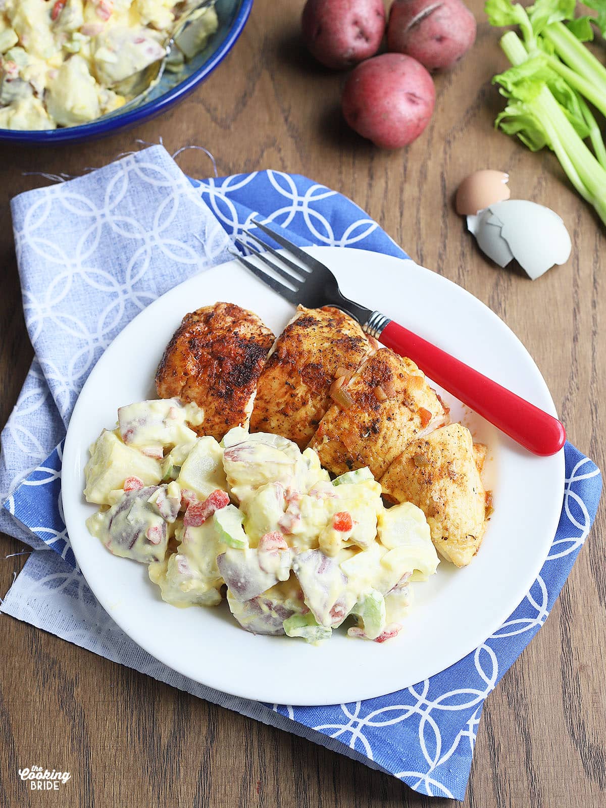 helping of potato salad on a white plate with a serving of roast chicken, a red fork and a blue and white napkin