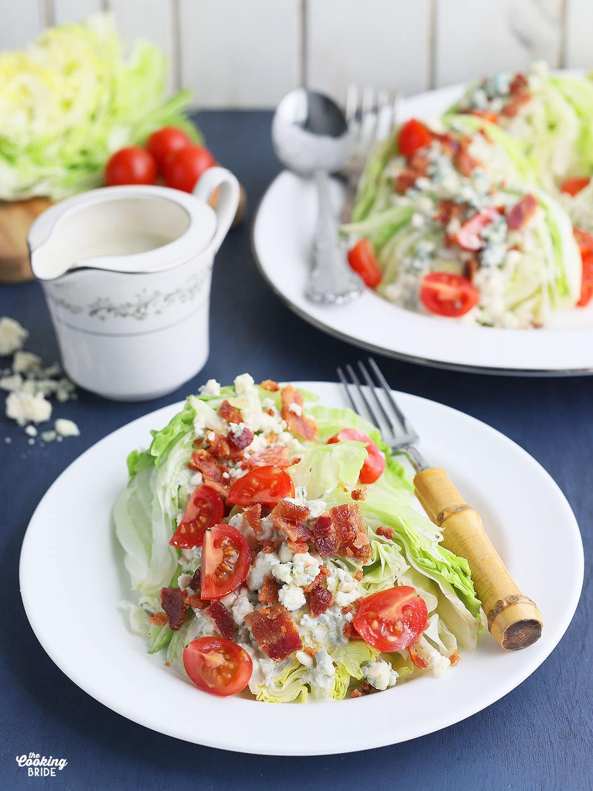 side view of a plated wedge salad with a small pitcher of dressing, platter of unserved salads and iceberg lettuce wedges in the background