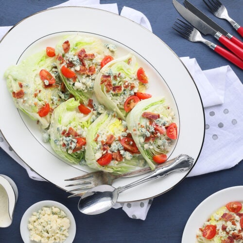 bacon and tomato wedge salads arranged on a white platter with extra blue cheese crumbles, dressing and a plated salad on the side