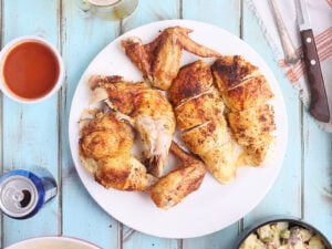 overhead shot of carved beer can chicken on a white plate with bowls of potato salad, cold beer and sauce on the side