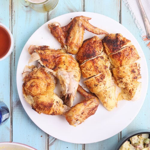 overhead shot of carved beer can chicken on a white plate with bowls of potato salad, cold beer and sauce on the side