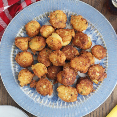 blue platter of squash puppies with a red and white checked napkin, iced tea, bowl of sauce and serving plate with one squash puppy to the side