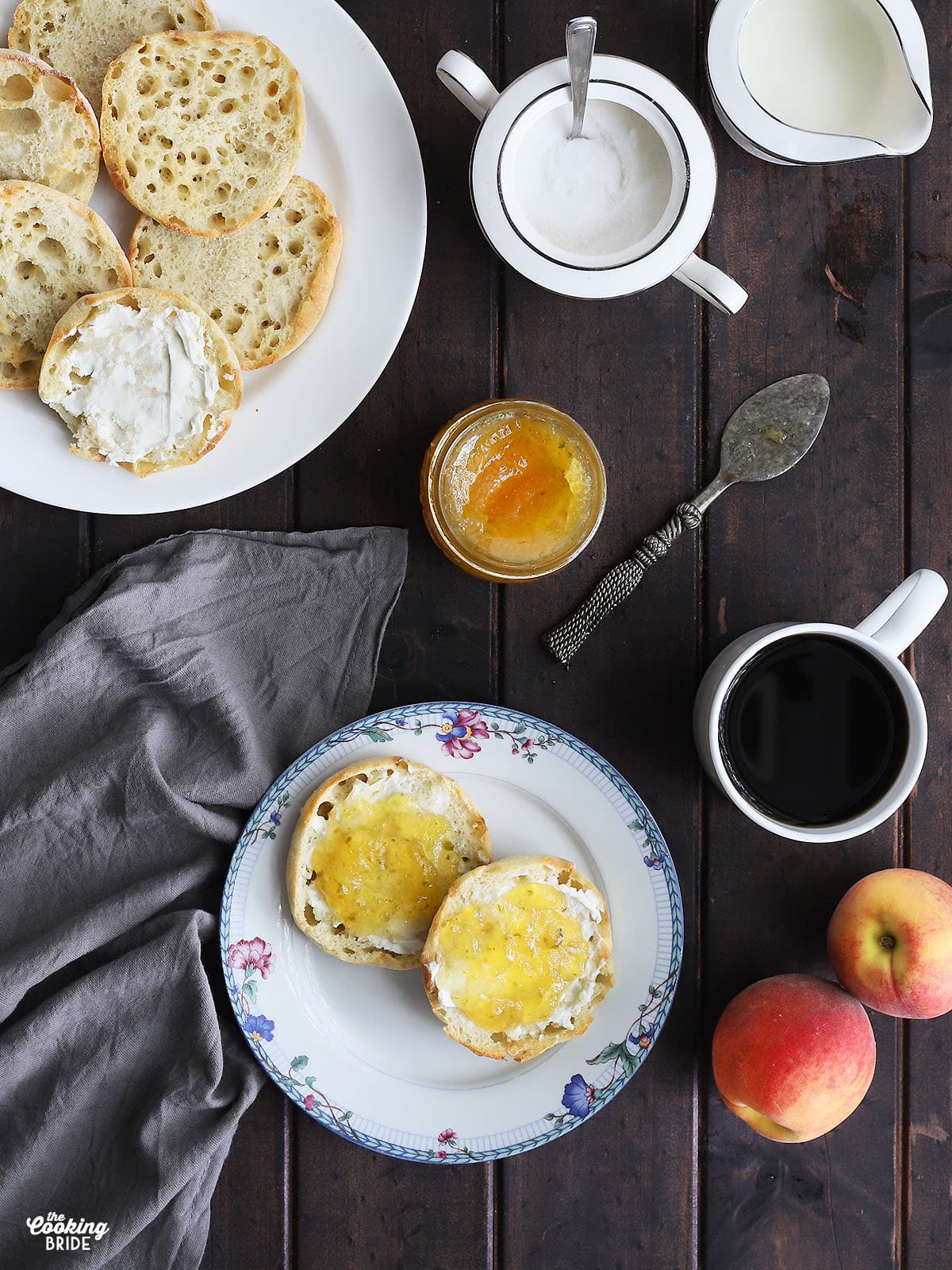 Two English muffins on a blue and white floral plate. Plate of English muffins, jam, coffee, peaches, cream and sugar to the side on a dark wooden table.