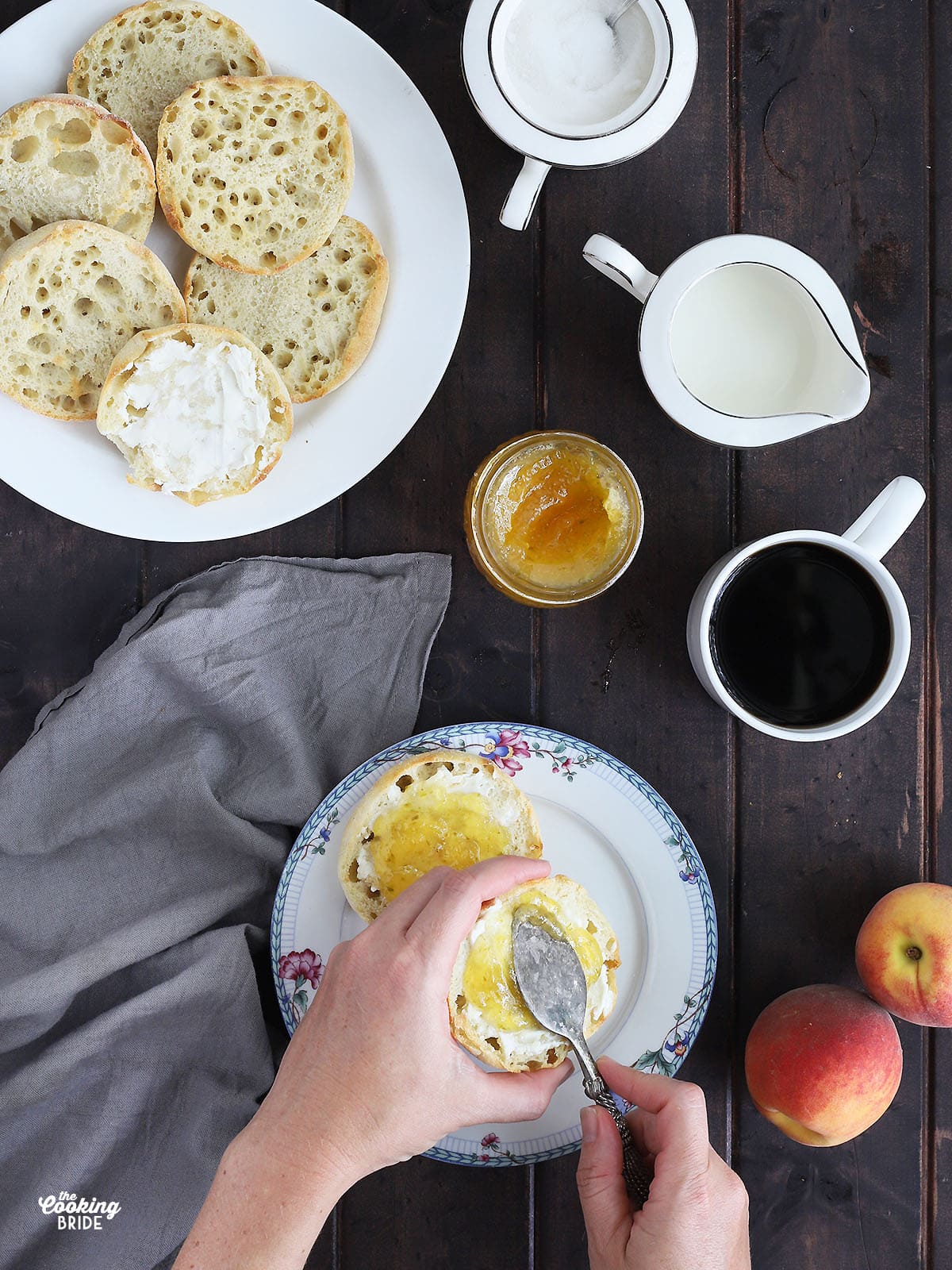 overhead shot of hands spreading peach jam over an English muffin. Plate of English muffins, jam, coffee, peaches, cream and sugar to the side on a dark wooden table.