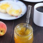 jar of peach jalapeno jam with the handle of a spreader sticking out. English muffin, mug of coffee and a peach in the background.