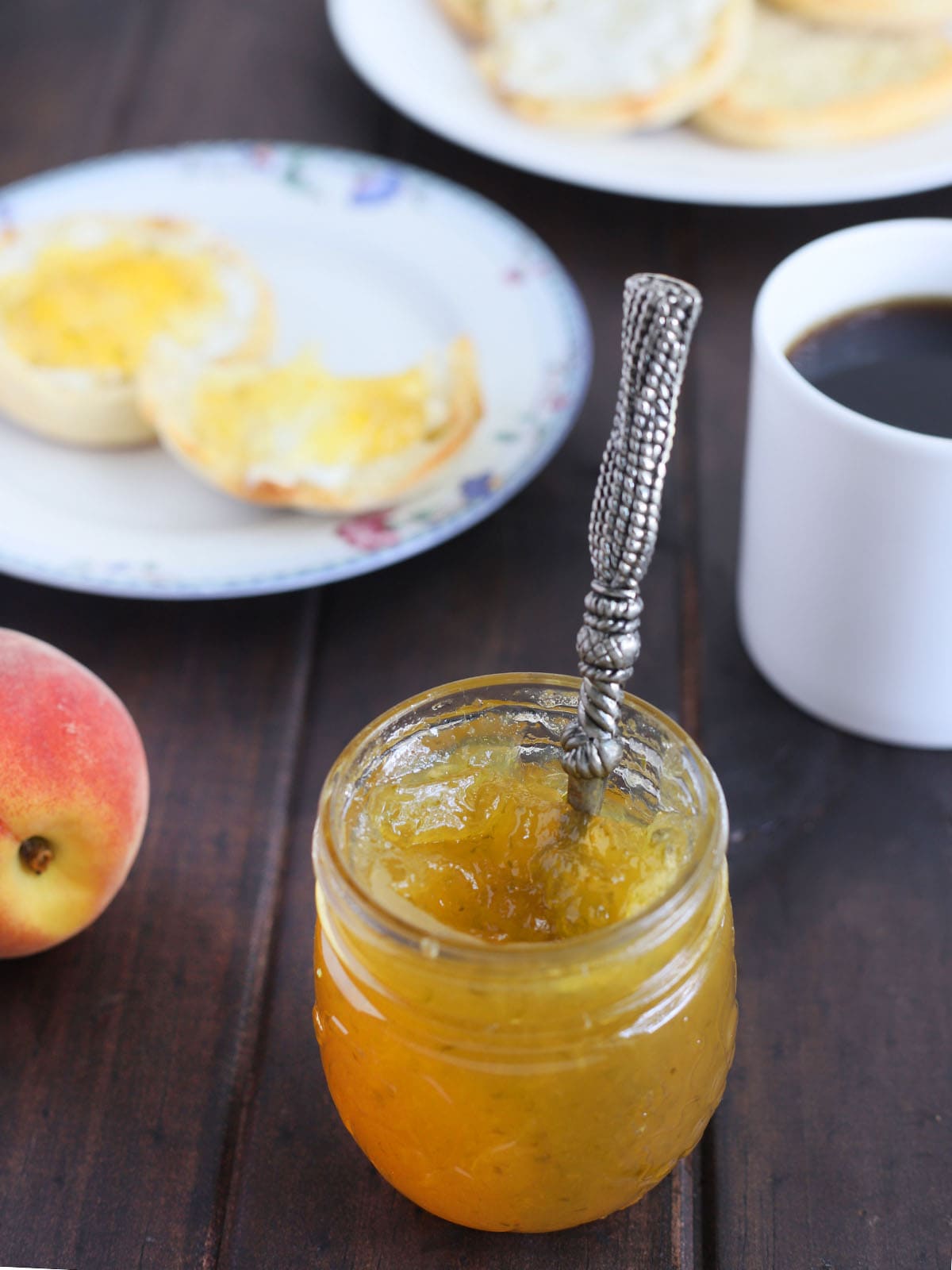 jar of peach jalapeno jam with the handle of a spreader sticking out. English muffin, mug of coffee and a peach in the background.