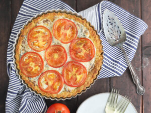 Baked southern tomato pie on a dark wooden table with fresh tomatoes, plates and forks to the side.