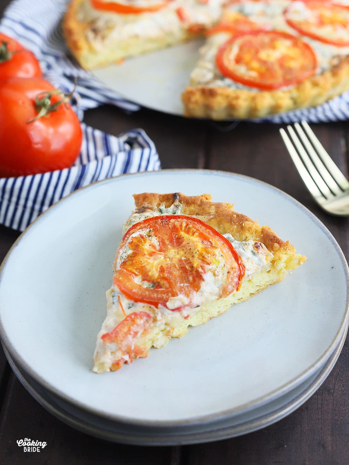 slice of Southern tomato pie on a grey plate with tomatoes and whole pie in the background