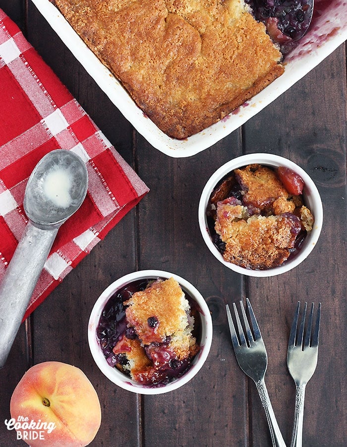 overhead shot of two bowls of blueberry peach cobbler. Pan of cobbler, two forks, ice cream scoop and fresh peach to the side.