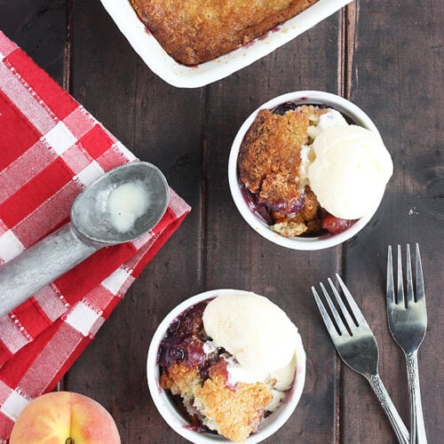 overhead shot of two bowls of blueberry peach cobbler with vanilla ice cream. Pan of cobbler, two forks, ice cream scoop and fresh peach to the side.