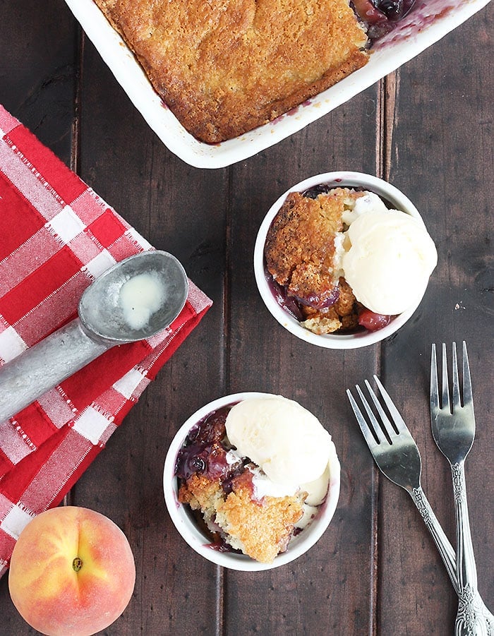 overhead shot of two bowls of blueberry peach cobbler with vanilla ice cream. Pan of cobbler, two forks, ice cream scoop and fresh peach to the side.