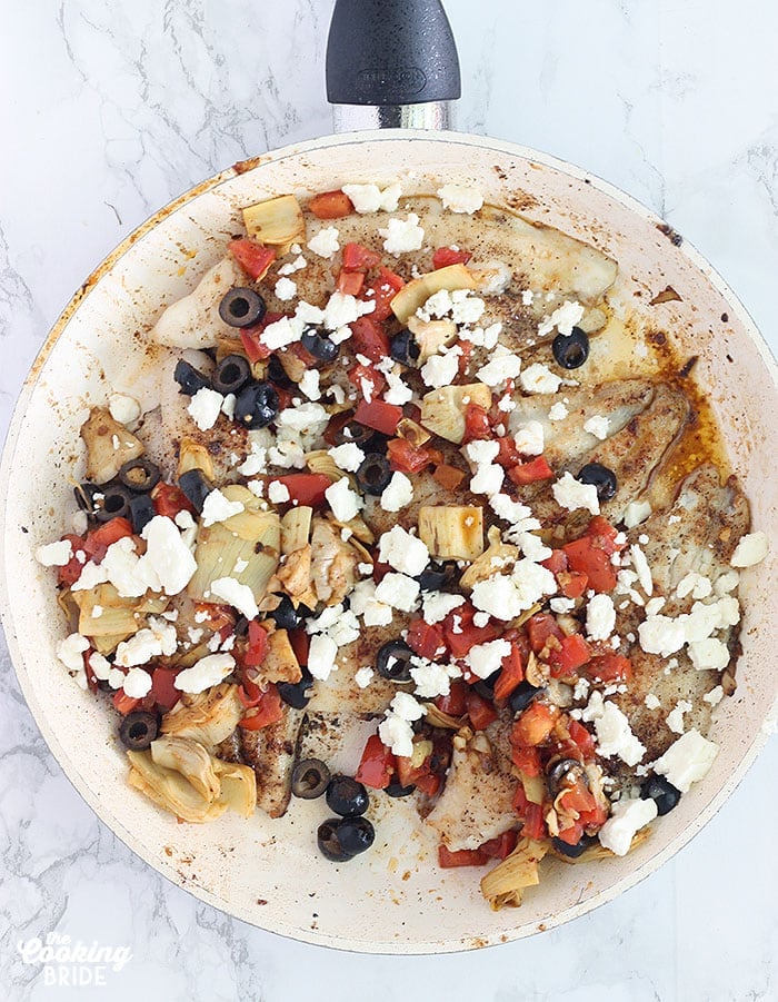 sauteed catfish fillets seasoned with salt, pepper and oregano in a skillet topped with artichoke hearts, feta, diced Roma tomatoes and black olives