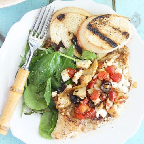Seasoned catfish fillets topped with artichoke hearts, black olives, tomatoes and feta cheese on a white plate with a fork and toasted bread