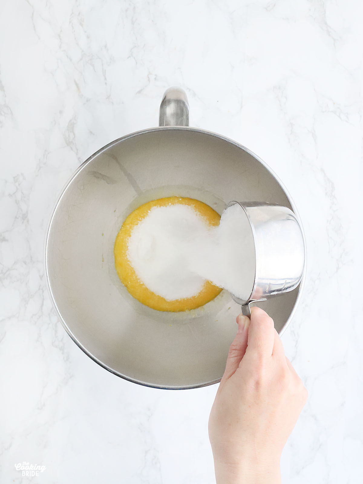 hand pouring sugar into a stainless steel mixing bowl with egg yolks