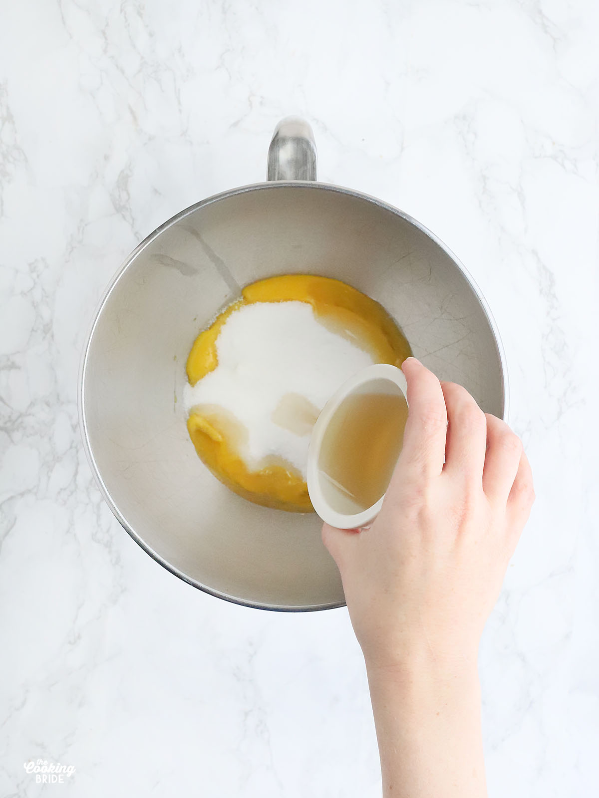 hand pouring spiced rum into a stainless steel mixing bowl with egg yolks and sugar