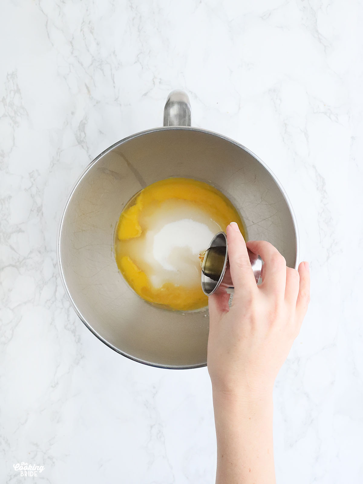 hand pouring vanilla extract into a stainless steel mixing bowl with egg yolks and sugar