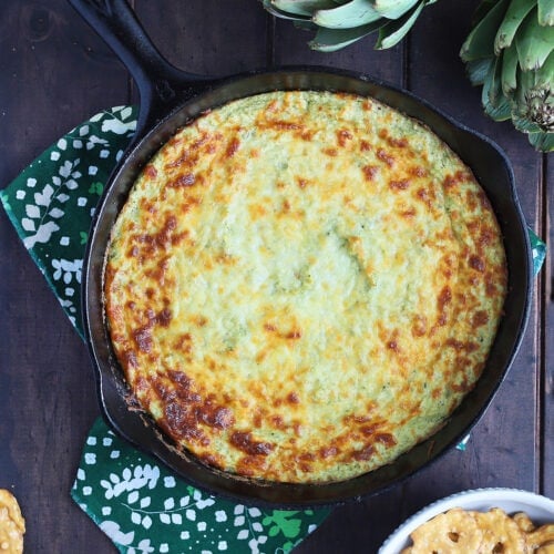 overhead shot of a cast iron skillet of hot artichoke dip with two whole artichokes and pretzel chips scattered to the side