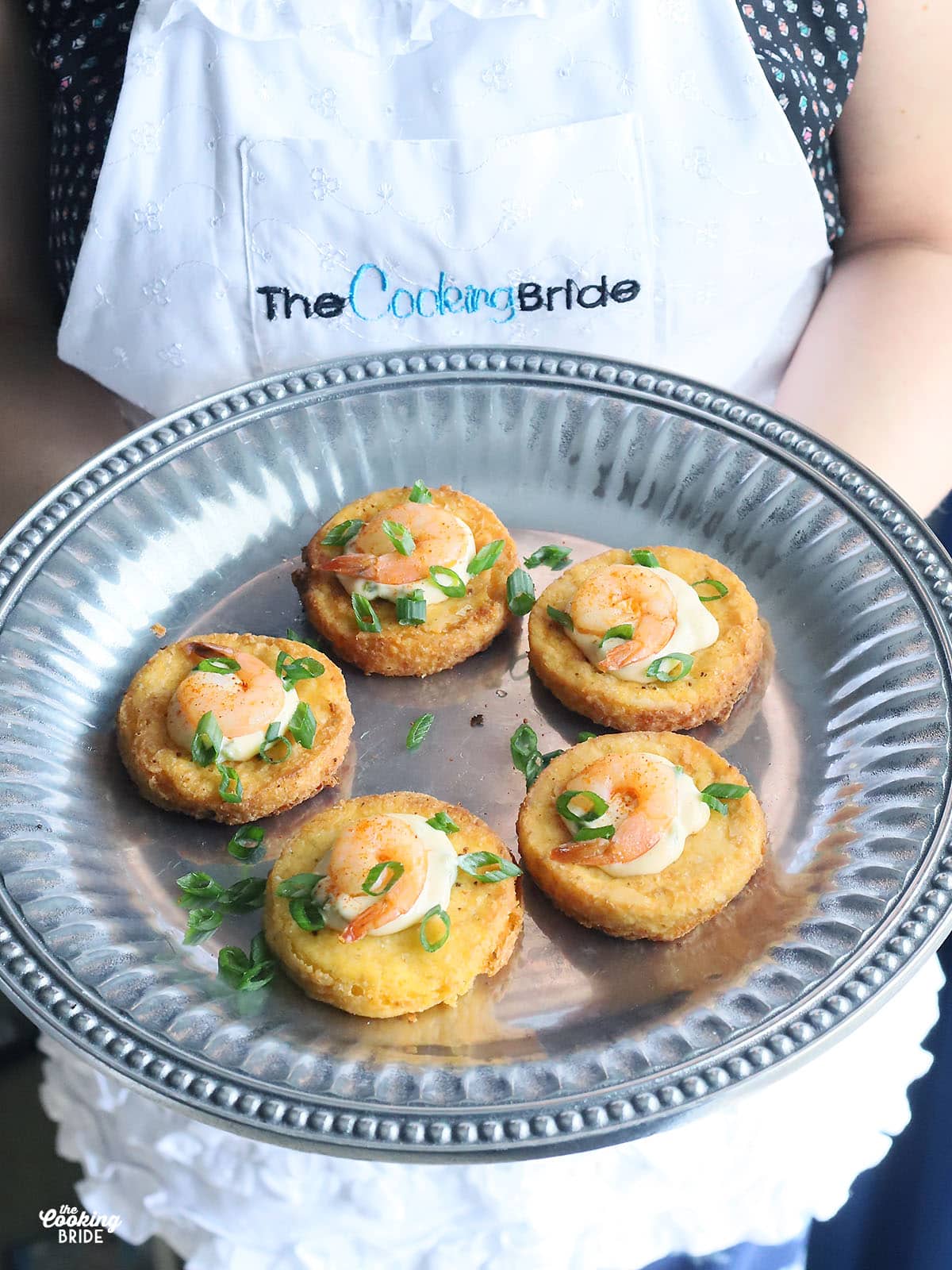person holding a pewter platter of five shrimp on fried grit cakes garnished with green onions