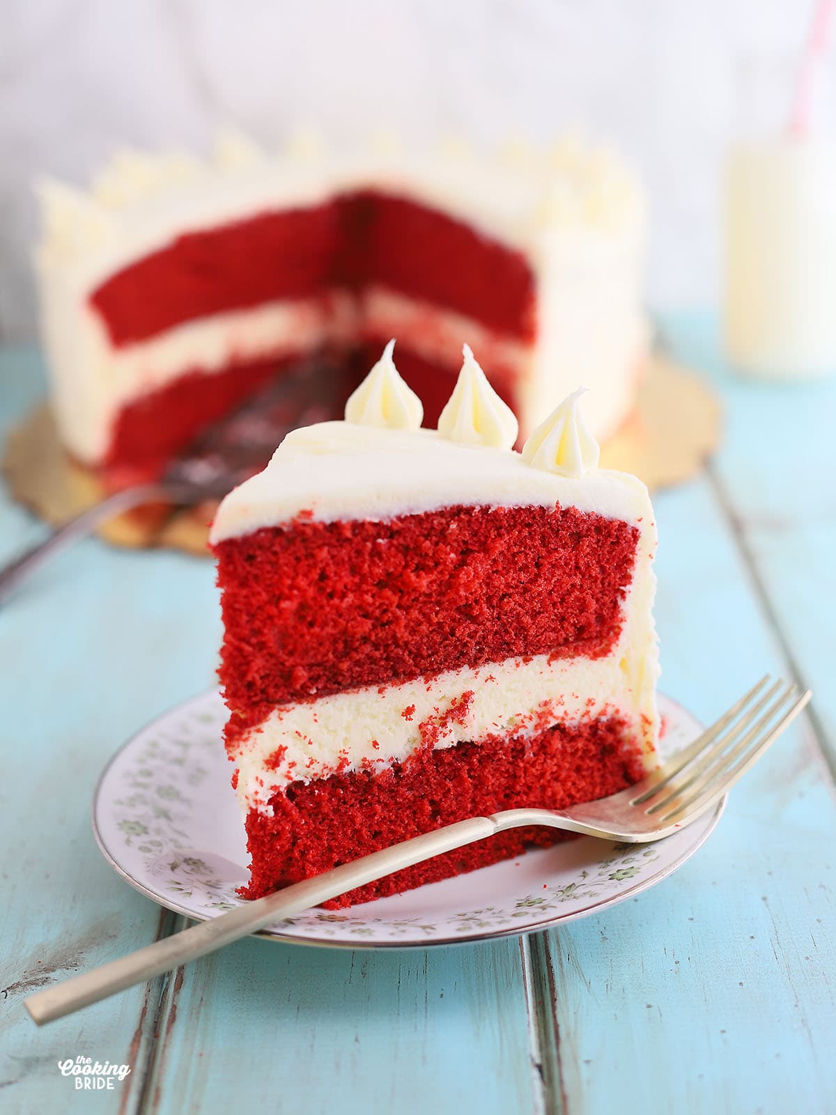 single slice of red velvet cake on a plate with a fork, cut cake in the background