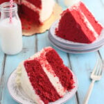 side shot of a slice of red velvet cake on a plate with a second slice, a bottle of milk and the remaining cake in the background