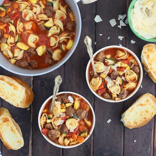 overhead shot of two white bowls of meatball tortellini soup with a pot of soup, parmesan cheese an sliced of cheese toast to the side