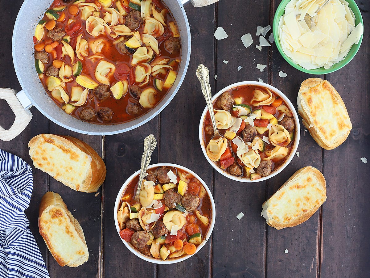 overhead shot of two white bowls of meatball tortellini soup with a pot of soup, parmesan cheese an sliced of cheese toast to the side