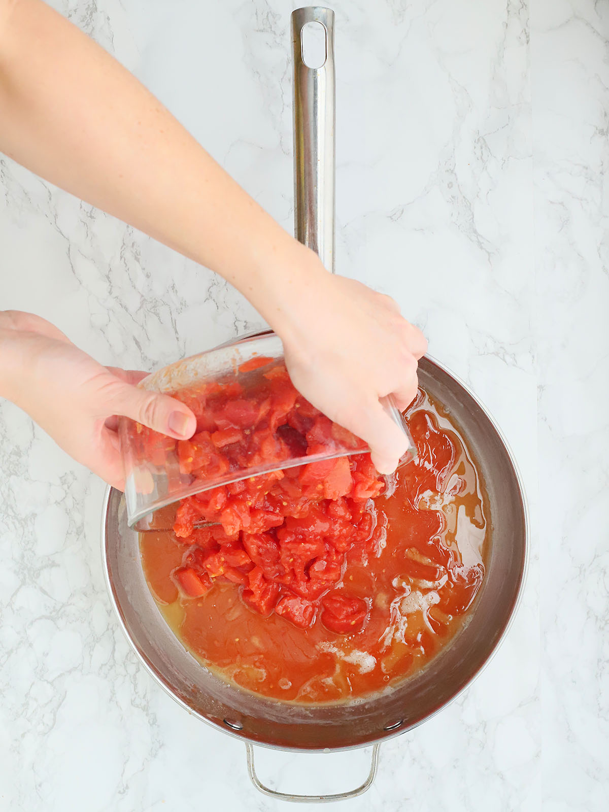 overhead shot of hands pouring diced tomatoes into a stainless steel skillet with flour and bacon grease