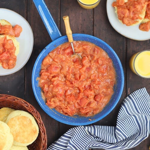 overhead shot of a blue skillet of tomato gravy with plates of biscuits topped with gravy and a basket of biscuits to the side on a dark wooden background