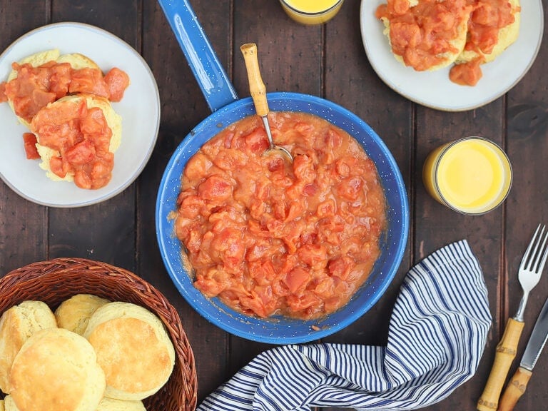 overhead shot of a blue skillet of tomato gravy with plates of biscuits topped with gravy and a basket of biscuits to the side on a dark wooden background