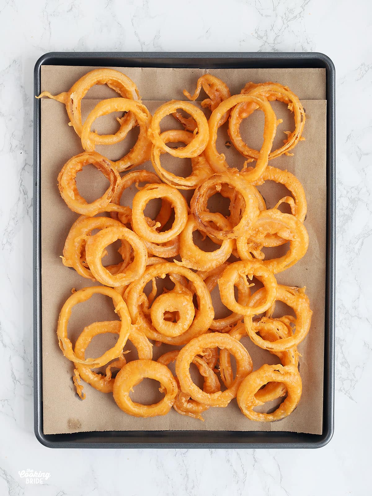 Overhead shot of beer battered onion rings draining on a cookie sheet lined with brown paper bags