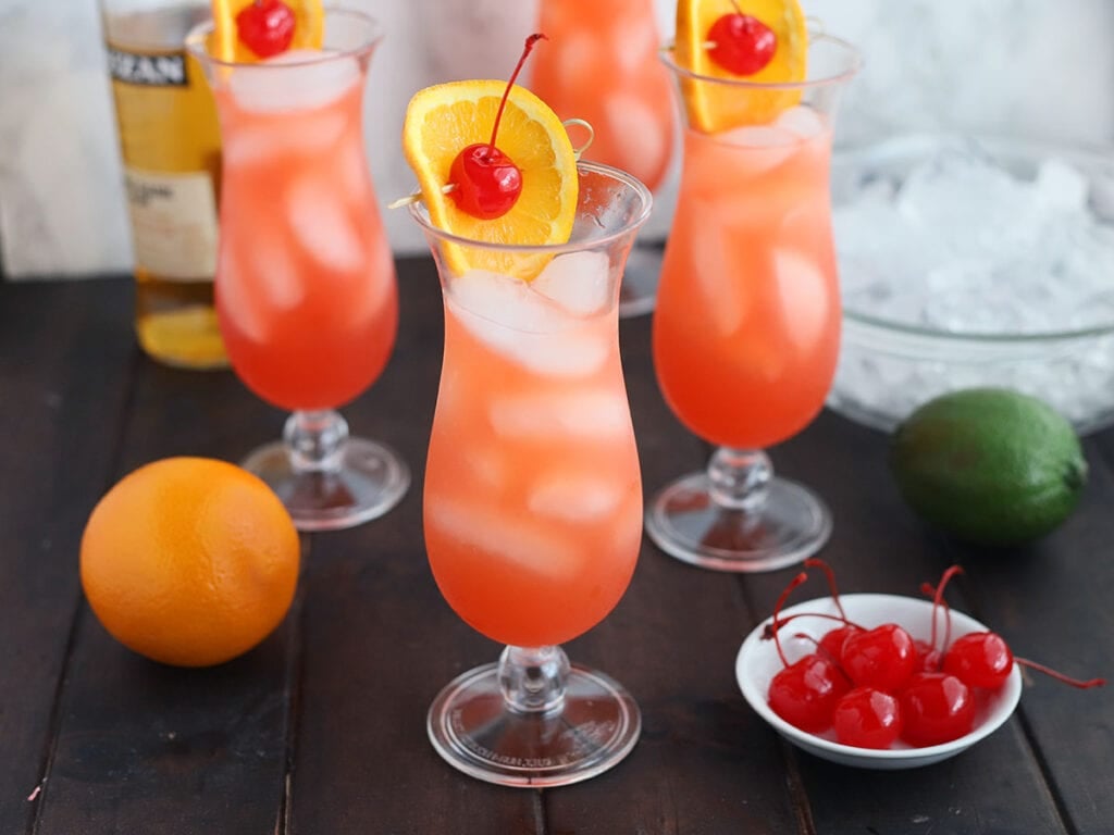 side shot of a hurricane cocktail garnished with a cherry and orange slice, three other glasses in the background