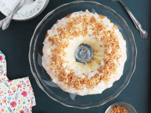 overhead shot of glazed Louisiana crunch cake on a glass cake pedestal with floral napkin, plates and dish of toasted coconut to the side