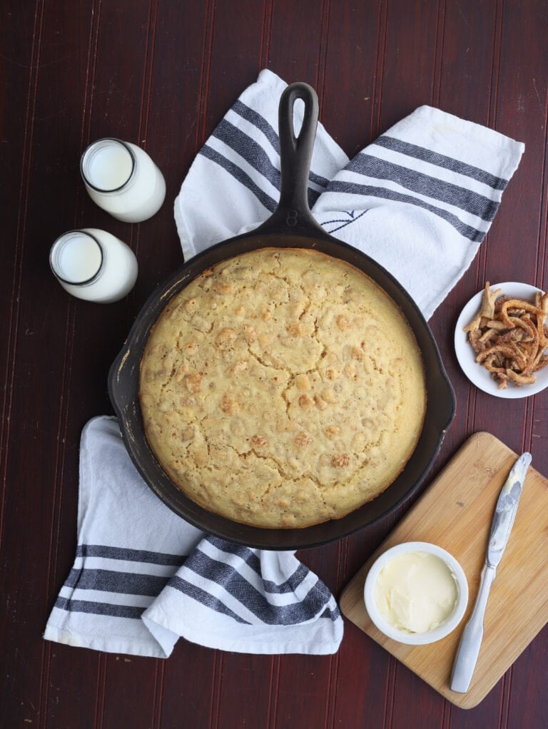 overhead shot of cracklin' corn bread in a cast iron skillet with butter, milk and a dish or cracklings to the side