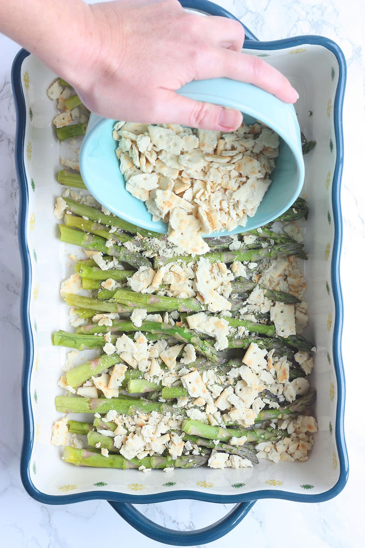 hand sprinkling crushed saltines over asparagus spears in a casserole dish